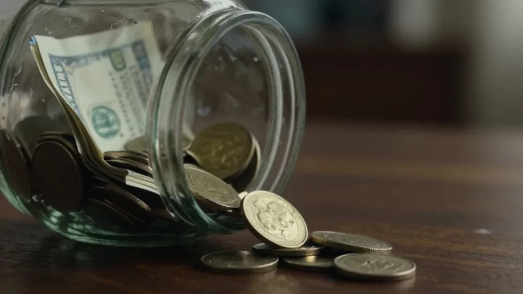 Monedas de oro que se desbordan de un frasco de vidrio agrietado sobre una mesa de madera.